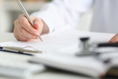 Female doctor hand hold silver pen filling patient history list at clipboard pad. Physical, exam, er, disease prevention, ward round, visit check, 911, prescribe remedy, healthy lifestyle conceptの写真素材
