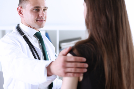 Handsome friendly male doctor talk with thankful female patient at office and greet her with hand on shoulder gesture. Welcoming friend, trust, physical, healthy lifestyle, exam interview conceptの写真素材