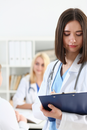 Beautiful smiling female doctor hold clipboard pad and fill something with silver pen. Physical, illness prevention, prescribe remedy, ward round, therapeutist assistance, healthy lifestyle conceptの写真素材
