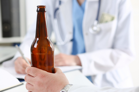 Alcoholic hold in hand empty bottle at doctor reception office closeup. Grief and despair, intoxicated, change life, drunkenness lifestyle, sobriety and temperance, bad habit dependence conceptの写真素材