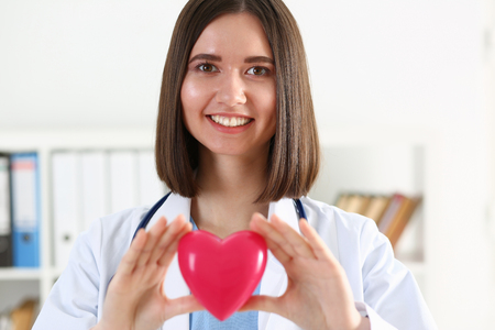 Female medicine doctor hold in hands red toy heart in front of her chest. Cardio therapeutist, student education, physician make cardiac physical, heart rate measure, arrhythmia conceptの写真素材