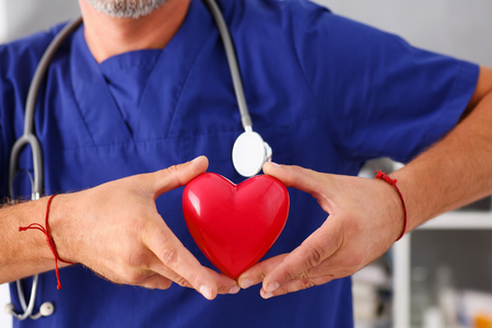 Male doctor wearing blue uniform hold in arms red toy heart closeup. Cardio therapeutist, student education, CPR, 911 life save, physician make cardiac physical, pulse rate measure, arrhythmiaの写真素材