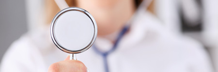 Female medicine doctor hand holding stethoscope head closeup in front of her chest. Medic shop or store, physical and disease prevention, er consultant, 911, pulse measure, healthy lifestyle conceptの写真素材