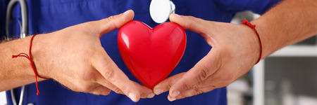 Male doctor wearing blue uniform hold in arms red toy heart closeup. Cardio therapeutist, student education, CPR, 911 life save, physician make cardiac physical, pulse rate measure, arrhythmiaの写真素材