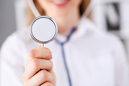 Female medicine doctor hand holding stethoscope head closeup in front of her chest. Medic shop or store, physical and disease prevention, er consultant, 911, pulse measure, healthy lifestyle conceptの写真素材