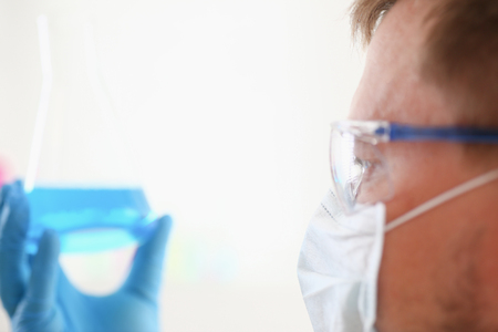 A portrait of a young surgeon chemist's doctor looks at a container with a blue liquid and a mask is fought with viruses and a vaccine for vaccines against diseasesの写真素材