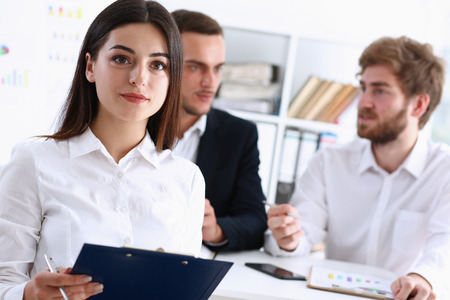 Beautiful smiling cheerful girl at workplace look in camera with colleagues group in background. White collar worker at workspace, job offer, modern lifestyle, client visit, profession train conceptの写真素材