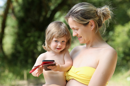 Happy little smiling child in the arms of the mother on a natural background. Holds a smartphone in the hands of watching developing video plays gamesの写真素材