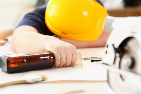 Arm of drunken worker in yellow helmet hold liquor bottle sleeping at table closeup. Manual job workplace, DIY inspiration, fix shop, hard hat, industrial education, profession careerの写真素材