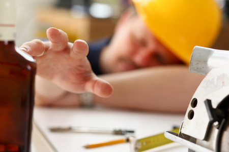 Arm of drunken worker in yellow helmet hold liquor bottle sleeping at table closeup. Manual job workplace, DIY inspiration, fix shop, hard hat, industrial education, profession careerの写真素材