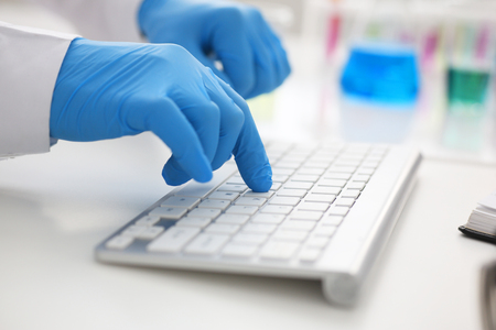 Employee chemical laboratory teacher chemist knocks his fingers keyboard makes notes electronic journal records the analysis data from reactions examining test tubes with the substance arm in glovesの写真素材