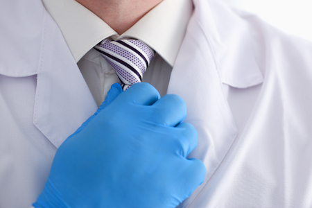 A male doctor in a dressing gown and shirt adjusts his tie with his hand in a blue gloveの写真素材