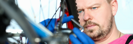 Male millennial repair man bicycle service in blue protective gloves adjusts bike to prepare for the...