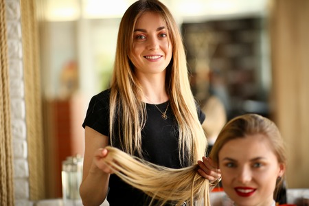 Young Hairdresser Holding Hair of Female Client. Smiling Hairstylist Picking Haircut for Girl. Two Blonde Woman in Beauty Salon. Stylist Beautician Styling Hairdo Looking at Camera Photographyの写真素材
