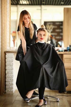Pretty Hairdresser and Girl with Wavy Hairstyle. Female Client with Curls and Bun Sitting in Beauty Studio. Woman with Curly Hair and Hairstylist with Long Straight Blonde Hair Vertical Photographyの写真素材
