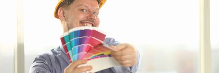 Portrait of Happy Male Designer Hold Color Palette. Architecture Draft Tools Colour Swatches for Selection. Freelance Joyful Foreman in Yellow Protective Helmet Posing with Paint Swatchの写真素材