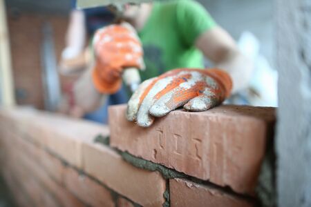 Close-up of big masonry made of red blocks. Builder man wearing special protective gloves and outfit...