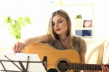 Woman Practice Acoustic Guitar Playing at Home. Female Player Performing while Sitting on Couch in Living Room. Blond Lady Turning Notes at Music Stand. Caucasian Girl Taking Chordsの写真素材