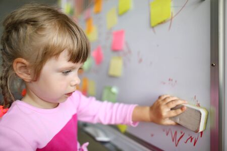 Portrait of smart small girl standing in modern classroom and holding special erasing marker utility...