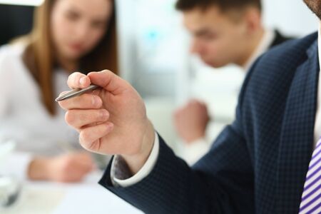 Focus on male hand holding writing pen and wearing classy suit and striped tie. Smart people talking about business project and strategy. Company meeting concept. Blurred backgroundの写真素材