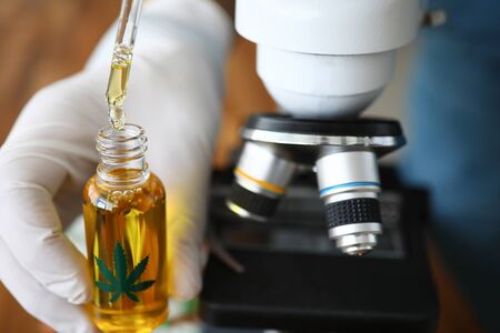 Focus on male hands in gloves holding flask with cannabinoid oil and dripping drops in glass bottle. Lab with microscope. Medicine and healthcare concept. Blurred backgroundの写真素材