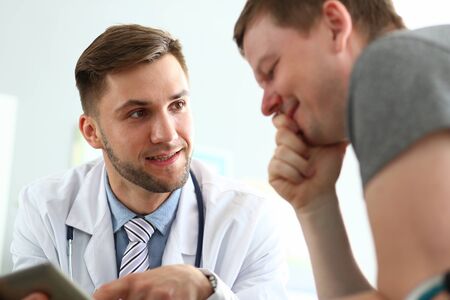 Portrait of smiling doctor reporting good news to cheerful man. Visitor looking to tablet with gladness. Medical treatment and health care concept. Blurred backgroundの写真素材