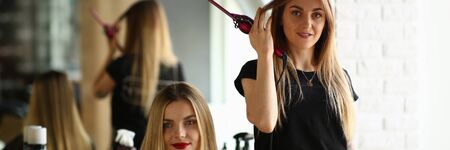 Young Girls Hairdresser and Beauty Salon Client. Hairstylist Holding Curling Iron in Hand. Female Customer Sitting in Studio for Making Haircut. Two Beautiful Blonde Woman. Styling Hairdoの写真素材