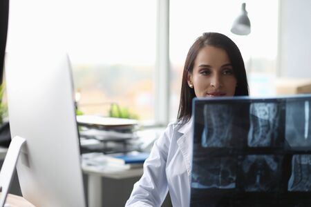 Portrait of smiling woman doc sitting at workplace and carefully examining at spine skiagram....