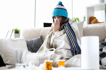 Portrait of of poor lady sitting in living room covered in blanket and suffering from common cold. Woman having severe flu at home indoors and sneezing into tissue. Hard virus conceptの写真素材