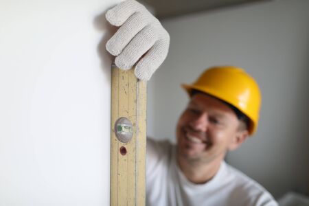 Focus on handyman hands holding measuring ruler and fixing straightness of wall. Builder wearing helmet and protective gloves. Person at house constructionの写真素材
