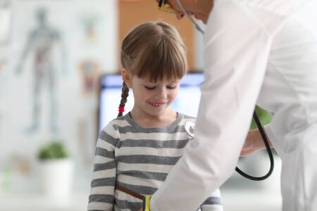 Portrait of joyful little female standing in office of modern hospital and looking down with interest. Professional female therapist checking patients condition. Health checkup conceptの写真素材