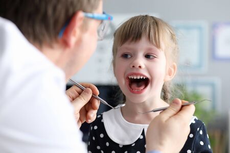 Male dentist look at open mouth litle happy girl child againlt hospital office background portrait....