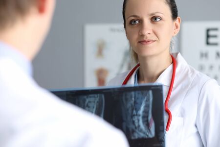 Portrait of cute female physician smiling and looking at colleague with gladness. Male doc holding...