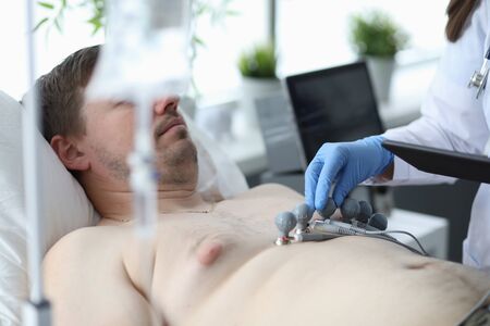 Portrait of sick male patient lying on medical couch. Smart female practitioner examining person and making cardiogram. Medicine and healthcare conceptの写真素材