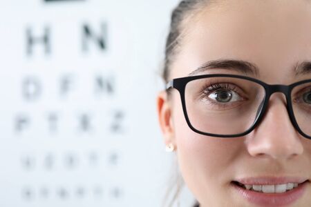 Focus on professional ophthalmologist looking at camera with happiness. Pretty woman wearing glasses in stylish black frame. Letters table for eyesight check on background. Vision test conceptの写真素材