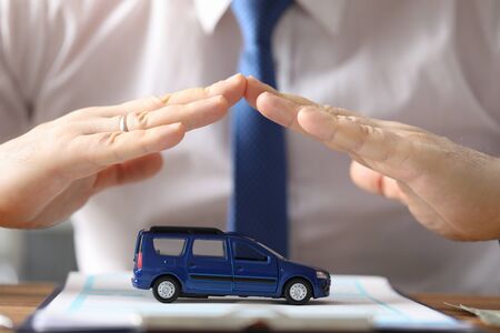 Close-up view of mans hands showing protection above blue car model. Businessman investing in transport. Presentable male in suit at office. Automobile insurance conceptの写真素材