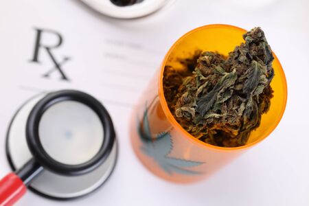 Close-up view of plastic orange transparent bottle filled with dry trimmed marijuana. Medical red stethoscope on recipe. Healthcare and folk medicine conceptの写真素材