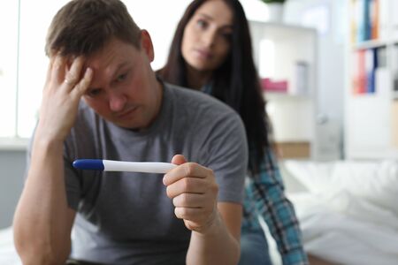 Portrait of anxious worried man holding pregnancy test in hand. Troublous female sitting behind and supporting male. People in cozy bedroom. Unexpected pregnancy conceptの写真素材