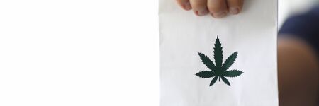 Focus on male hand holding white small paper bag with green cannabis symbol on it. Healthcare and medicines concept. Copy space in left side. Blurred backgroundの写真素材