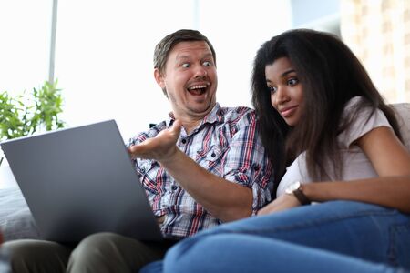 Portrait of surprised man showing something to african-american woman on laptop. Astonished female sitting next to husband on couch. Mixed race family conceptの写真素材