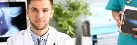 Portrait of handsome doctor working in modern clinic with colleagues. Young physician in white lab coat and blue shirt looking in camera with joy. Medical treatment conceptの写真素材