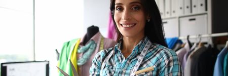 Portrait of smiling female tailor standing near comfortable workplace and looking at camera with...