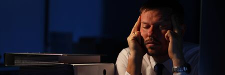 Portrait of man realizing amount of biz. Exhausted businessman sitting indoors and holding aching head with hands. Worker getting bogged down in work. Night at office conceptの写真素材