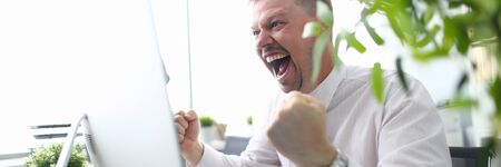 Portrait of cheerful man looking at computer monitor with gladness and making successful gesture. Businessman finishing project and wins big. Blurred backgroundの写真素材