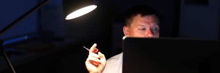 Portrait of working businessman sitting indoors and holding pen. Concerned business person looking at screen of modern high-tech laptop. Night at office conceptの写真素材