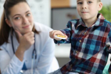 Focus on plate of yellow pills in hands of small joyful kid. Boy sitting in family doc office and looking at camera with joy. Pediatrician sitting near little child. Pediatrics and healthcare conceptの写真素材