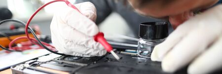 Close-up of soldering laptop parts in computer service or gadget support center. Technician male holding tool and examining circuit board looking through small magnifying glassの写真素材