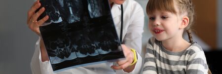 Portrait of smart female doctor sitting in modern clinic office and showing x-ray shot to little...