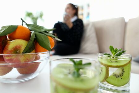 Ecological natural fruits on table and in drinks. In background, girl is sitting on sofa, waiting at her feet, image blurry. On table vase are tangerines and apples. Glasses drink with kiwi slices.の写真素材