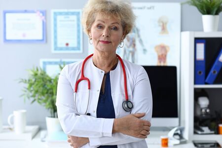 Experienced female doctor ready receive patients. An elderly woman doctor stands in white coat with stethoscope on her neck. Interior therapist office visual aids on wall modern furniture.の写真素材